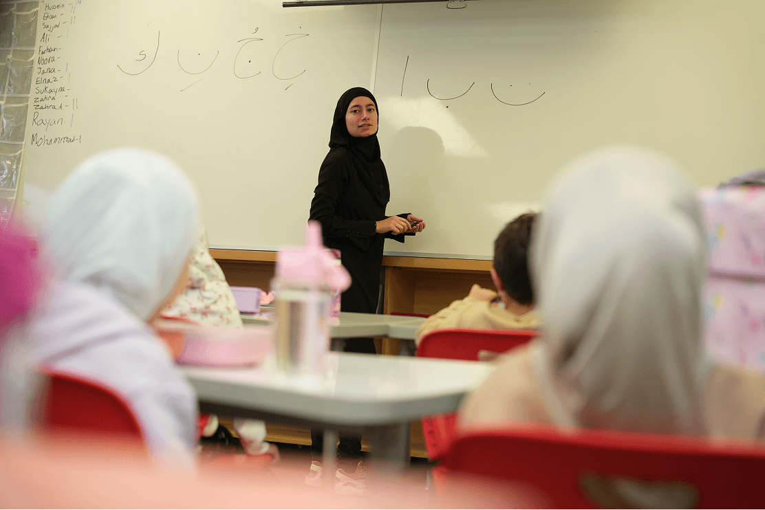 Female teacher wearing a hijab writing Arabic letters on a whiteboard in a classroom with students.