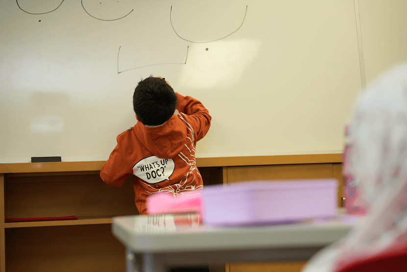 Child in an orange hoodie drawing smiley faces on a whiteboard in a classroom.