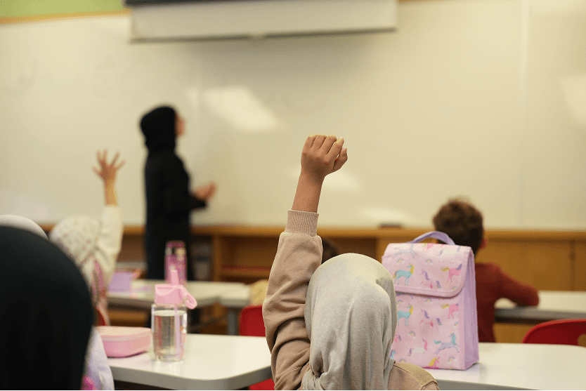Students sitting at desks in a classroom, with two raising their hands, and a teacher standing near a whiteboard.