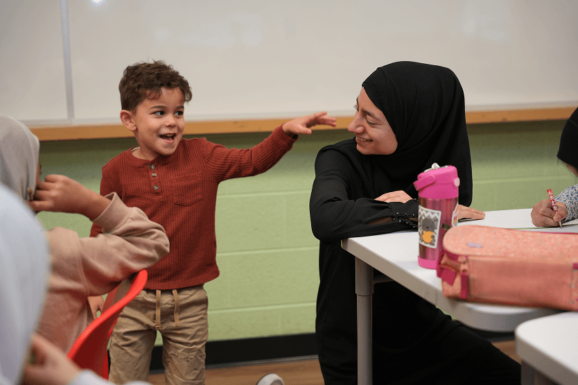 Young boy in a red sweater smiling and reaching out to a woman in a black hijab who is seated at a desk and smiling back.