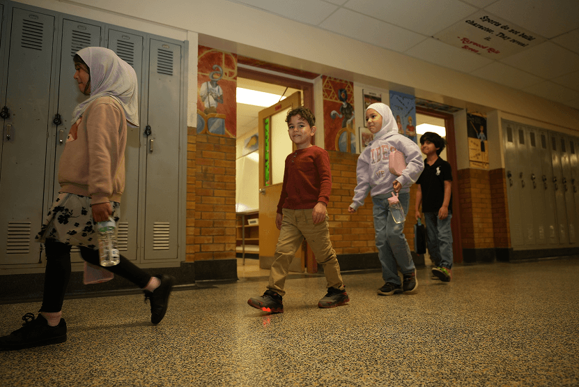 Four children walking in a school hallway past lockers and decorated walls, two girls wearing hijabs and one boy wearing light-up shoes.