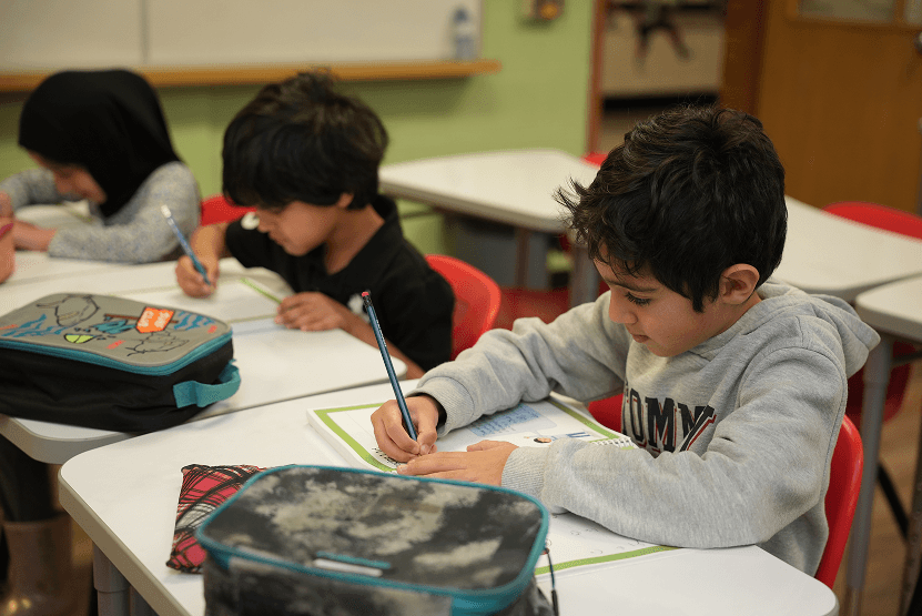 Three young students seated at desks in a classroom, focused on writing in their notebooks.