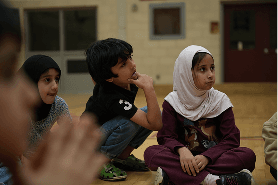 Three children sitting on the ground, one girl wearing a white hijab looking to the side, a boy crouching with his hand on his chin, and another girl wearing a black headscarf.