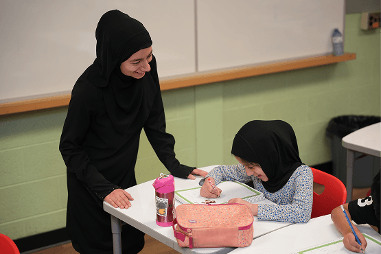 A female teacher in a black hijab smiling and leaning over to assist a young girl in a black hijab who is drawing at a desk with a pink pencil case and water bottle.